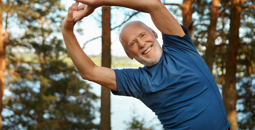 Outdoor shot of happy energetic senior retired man enjoying physical training in park, doing side bends exercise, holding hands together, looking at camera with broad smile, warming up body before run