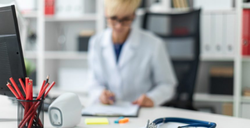 On the table is a phonendoscope. A beautiful young girl in a white robe working in a bright office at a computer Desk. She is holding a pen and a document. The girl has short white hair and wearing glasses. photo with depth of field