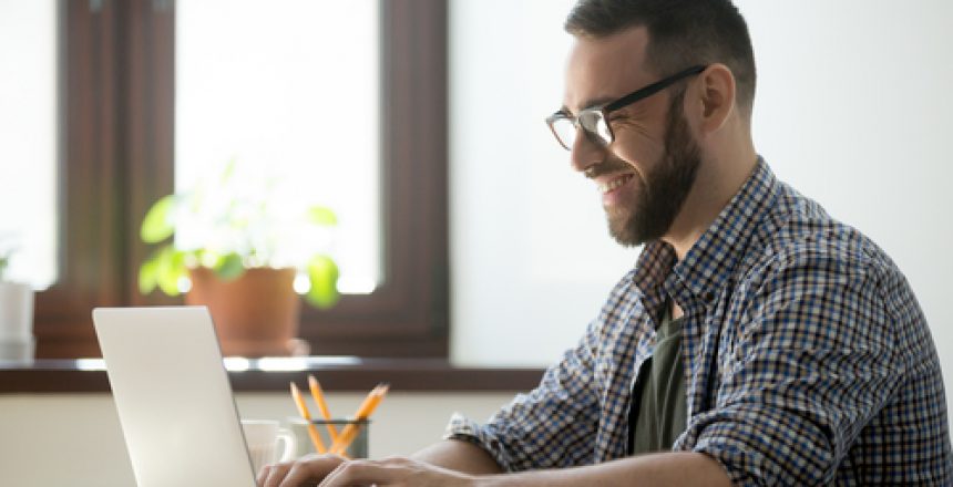 Happy male office worker smiling typing message at laptop, chatting with friends, writing positive email or consulting client online. Concept of laugher, positive work atmosphere, help and assistance